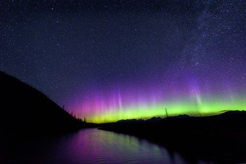 A stunning night sky filled with vibrant auroras reflecting in a calm river, surrounded by mountains and stars.