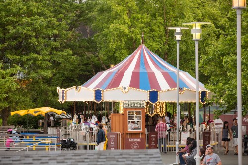A colorful carousel with a striped canopy, surrounded by trees and people enjoying a park setting.
