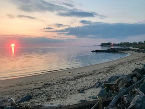 A serene beach at sunset, with calm waters reflecting the sun and a distant lighthouse on the shore.