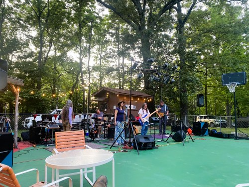 A live band performs outdoors on a colorful stage surrounded by trees, with a basketball hoop in the background.