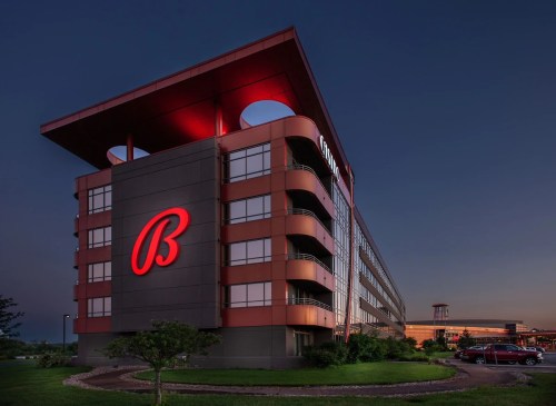 A modern hotel building at dusk, featuring a prominent red "B" logo and illuminated architectural details.