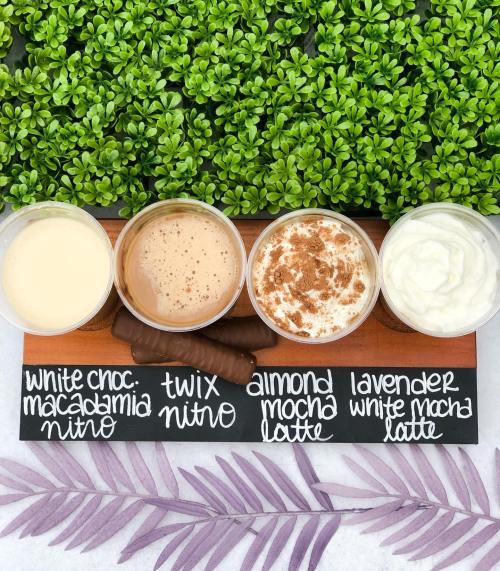 Four coffee drinks in clear cups on a wooden board, labeled with flavors, against a backdrop of greenery.