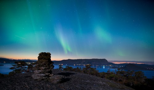 A serene landscape featuring a stone cairn under a vibrant aurora borealis and starry night sky over a calm lake.