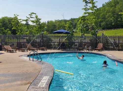 Two people play in a sunny pool surrounded by lounge chairs and greenery, with a shaded area in the background.