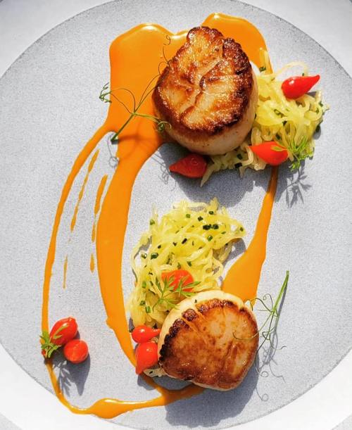 Two seared scallops on a gray plate, garnished with yellow squash, cherry tomatoes, and a vibrant orange sauce.