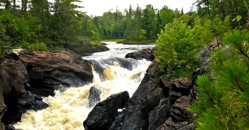 A rushing waterfall cascades over rocky terrain, surrounded by lush green trees and a serene river landscape.