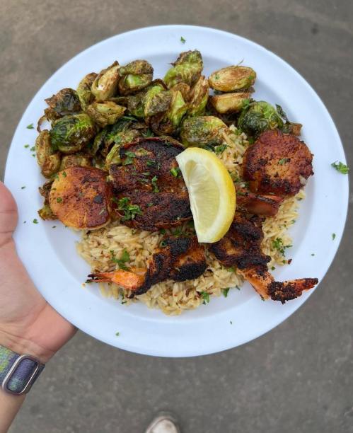 A plate of grilled shrimp and chicken on rice, served with roasted Brussels sprouts and a lemon wedge.
