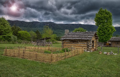 A rustic log cabin surrounded by a woven fence and a garden, set against a backdrop of dark, stormy clouds and mountains.