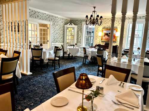 Elegant dining room with white tablecloths, floral wallpaper, and soft lighting, featuring empty tables and a chandelier.