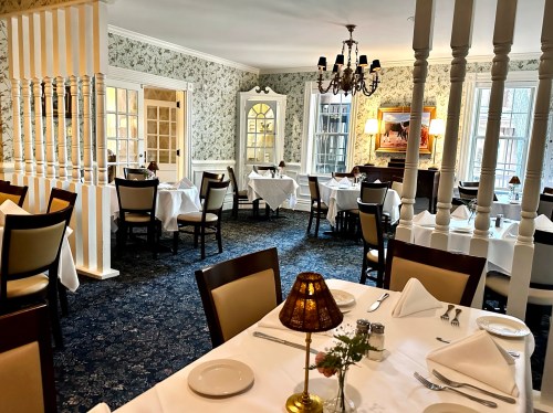 Elegant dining room with white tablecloths, floral wallpaper, and soft lighting, featuring empty tables and a chandelier.