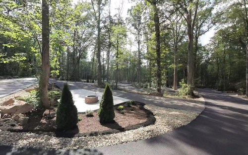 A scenic view of a forested area with a circular pathway, landscaped stones, and a fire pit surrounded by trees.