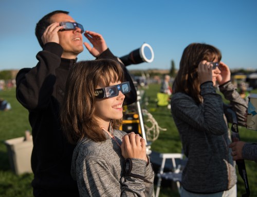 A man and two children wearing eclipse glasses, looking up at the sky during a solar eclipse event.