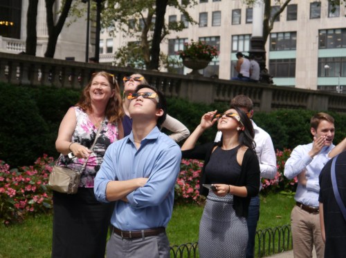 A group of people wearing sunglasses look up at the sky, surrounded by flowers and buildings in a park.