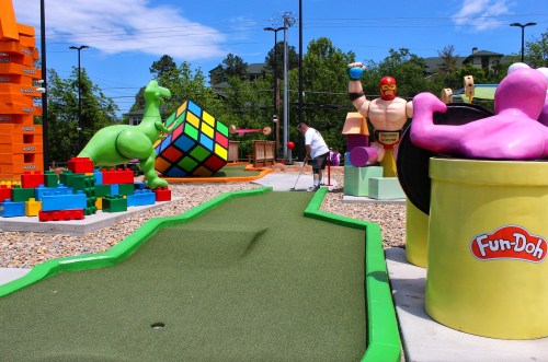 A colorful mini-golf course featuring a dinosaur, a giant Rubik's Cube, and playful sculptures, with a player in the background.