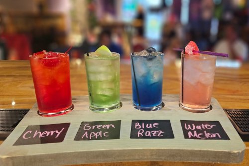 Four colorful cocktails on a wooden tray, labeled Cherry, Green Apple, Blue Razz, and Watermelon.
