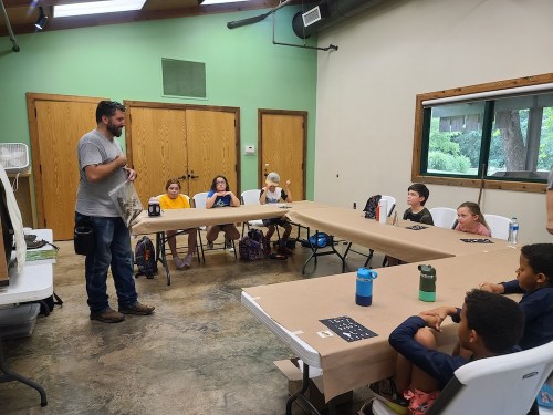 A group of children sits at tables while an adult speaks in a classroom setting.