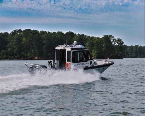 A state trooper boat speeds across a lake, creating waves, with trees in the background under a blue sky.