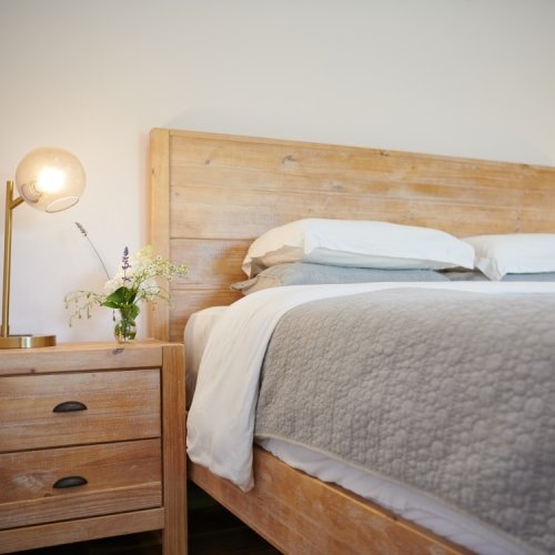 A cozy bedroom featuring a wooden bed frame, white bedding, a bedside table, and a lamp with a small flower vase.