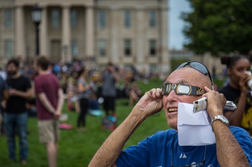 An elderly man adjusts his eclipse glasses, smiling, with a crowd and a historic building in the background.