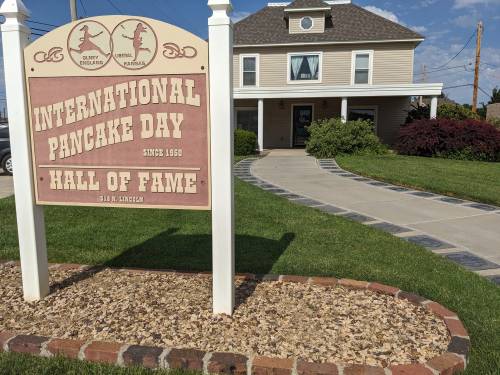 Sign for the International Pancake Day Hall of Fame, featuring a pathway and a large house in the background.
