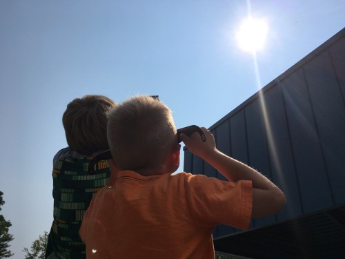 Two children looking up at the sun, one holding a device, with a clear blue sky in the background.