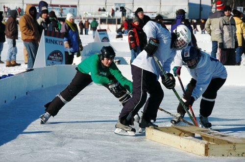 Three players compete in a snowy outdoor hockey game, with spectators in the background.
