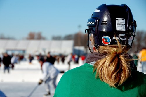 A person in a hockey helmet and green shirt watches a game on an outdoor rink, with spectators in the background.