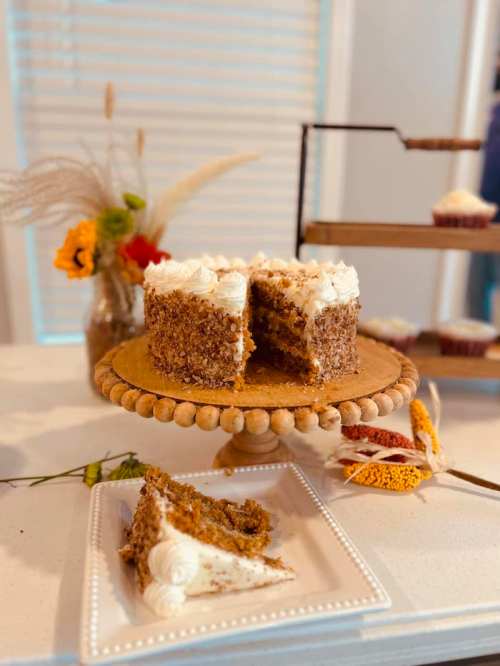 A layered carrot cake with cream cheese frosting, displayed on a wooden cake stand, with a slice on a plate nearby.