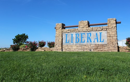 Sign reading "LIBERAL" in blue letters on a stone structure, surrounded by green grass and flowering bushes under a clear sky.