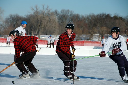 A group of hockey players in plaid jerseys skating on an outdoor rink, with trees in the background and a clear sky.