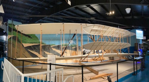 A large replica of a historic airplane displayed in a museum, with a scenic mural in the background.