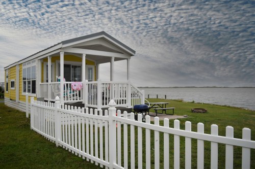 A yellow cottage by the water, featuring a white picket fence and a picnic area under a cloudy sky.
