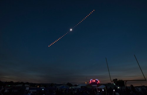 A time-lapse of a solar eclipse, showing the moon's phases against a dark sky, with a crowd and scoreboard below.