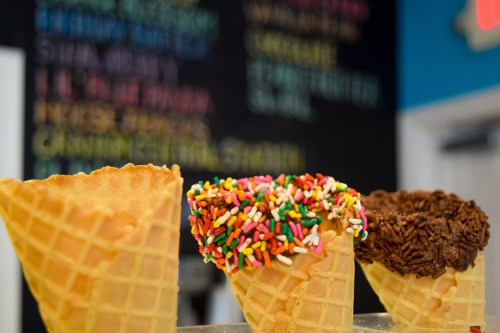 Three ice cream cones: one with colorful sprinkles, one with chocolate sprinkles, and one plain, against a colorful menu backdrop.