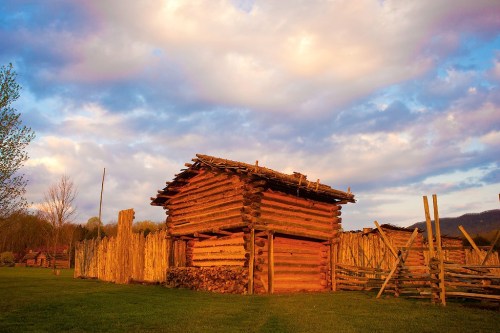A rustic wooden cabin surrounded by a wooden fence, set against a colorful sky at sunset.