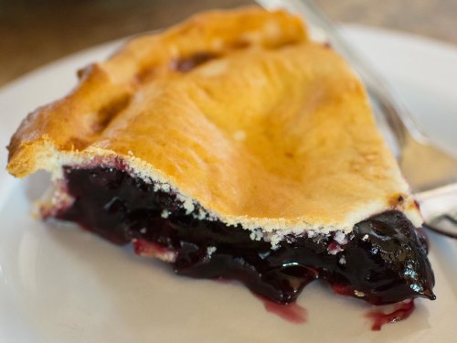 A slice of berry pie with a golden crust, revealing a rich, dark filling on a white plate.