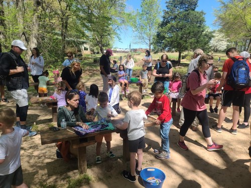 A lively outdoor scene with children and adults engaged in crafts at a picnic area surrounded by trees.