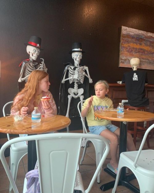 Two girls sit at a table in a café, enjoying snacks, with skeletons in hats seated nearby.