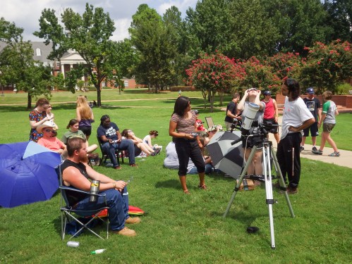 A group of people gathered outdoors, some sitting on chairs, observing through a telescope on a sunny day.