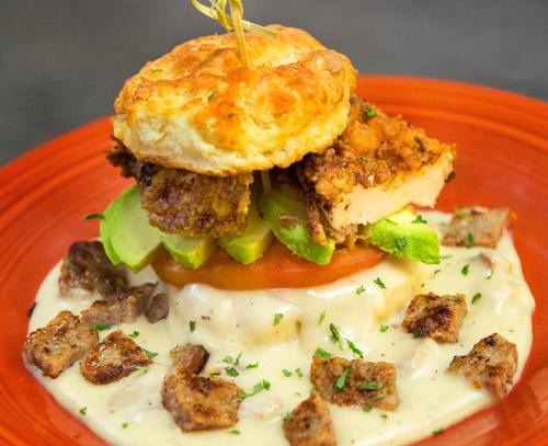 A biscuit sandwich with fried chicken, avocado, and tomato, served on creamy gravy with diced sausage on a red plate.