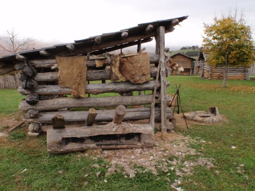 A rustic log structure with animal hides hanging, set in a grassy area with trees and additional buildings in the background.