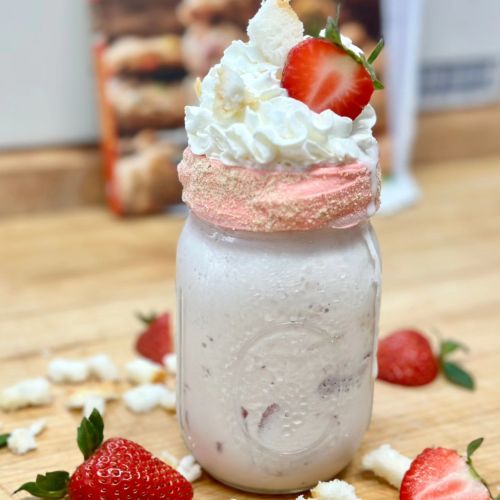 A creamy milkshake in a jar topped with whipped cream, a pink rim, and fresh strawberries, with cake pieces scattered around.
