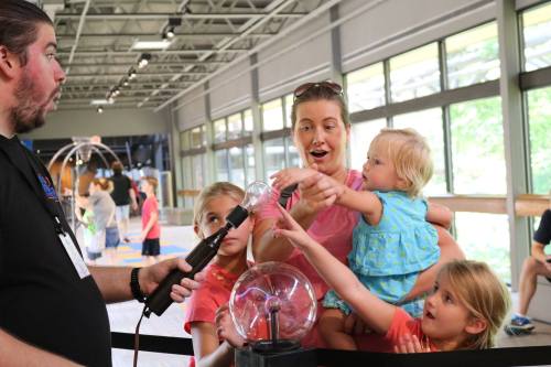 A man demonstrates a science experiment to a group of excited children and a woman in a bright, open space.