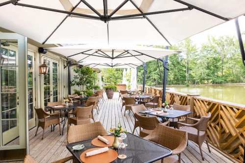 A scenic outdoor dining area with tables under large umbrellas, surrounded by greenery and a calm water view.
