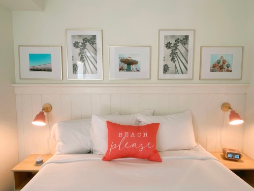 A cozy bedroom with a white bed, decorative pillows, and framed beach-themed artwork on the wall above.
