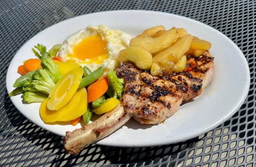 Grilled pork chop topped with apples, served with a sunny-side-up egg and a side of mixed vegetables.