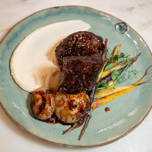 A plated dish featuring tender beef, caramelized onions, colorful roasted carrots, and a creamy puree on a blue plate.