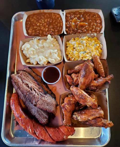 A metal tray filled with barbecue meats, including brisket, sausage, and wings, alongside sides of mac and cheese, beans, and corn.