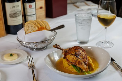 A plated dish featuring roasted chicken on a bed of creamy sauce, accompanied by bread, wine, and water on a dining table.