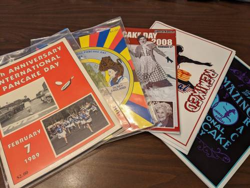 A collection of colorful pamphlets and flyers for International Pancake Day events from various years.